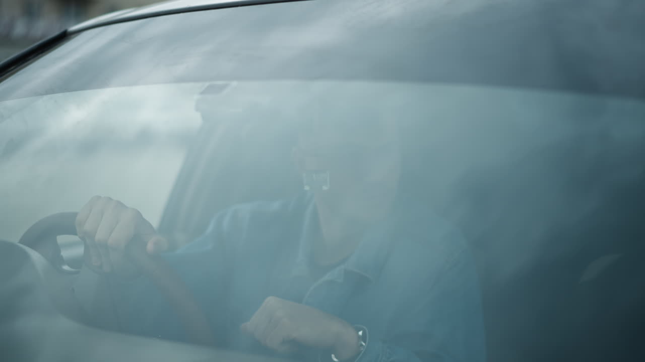 uomo bianco alla guida con le mani sul volante dietro un parabrezza appannato, espressione pensierosa, riflesso sfocato sul vetro, strada urbana visibile attraverso la foschia, dettaglio interno che mostra il cruscotto e le mani