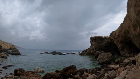 Sea-bay-with-rocks,-caves-and-cliffs-below-gray-cloudy-sky,-Malta