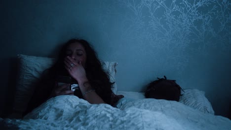 mujer se desplaza a través de las redes sociales y bosteza mientras está sentada en la cama por la noche