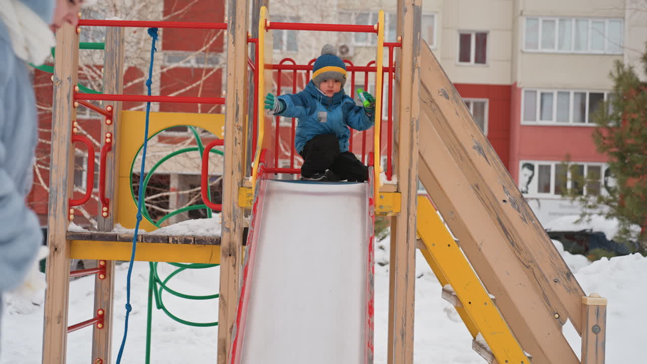 Son sliding down snowy playground slide mom watches nearby on cold winter day colorful play structure in urban apartment courtyard child in blue jacket and hat joyful supervised outdoor play family