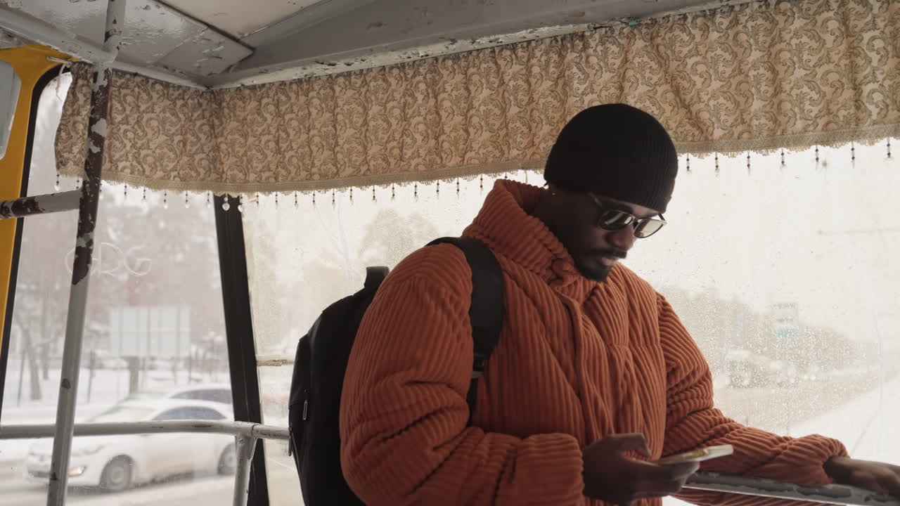 Uomo di colore che sale su un autobus giallo su una strada innevata, scavalca il marciapiede tenendosi al corrimano, con lo zaino in spalla, controlla il telefono, finestrini ghiacciati e traffico cittadino all'esterno, scena di transito urbano spontanea.