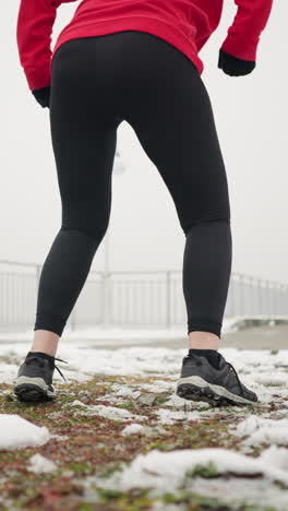 lady performing jump squats during winter outdoor workout on snowy ground wearing red hoodie black gloves amid foggy atmosphere with light poles iron railing sign post hill trees breath