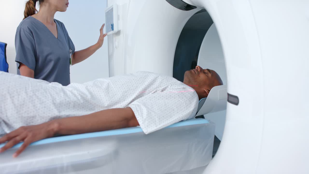 Tecnico asiatico che indossa una divisa grigia mentre utilizza un pannello per spostare un uomo afroamericano nel portale della TC per una scansione