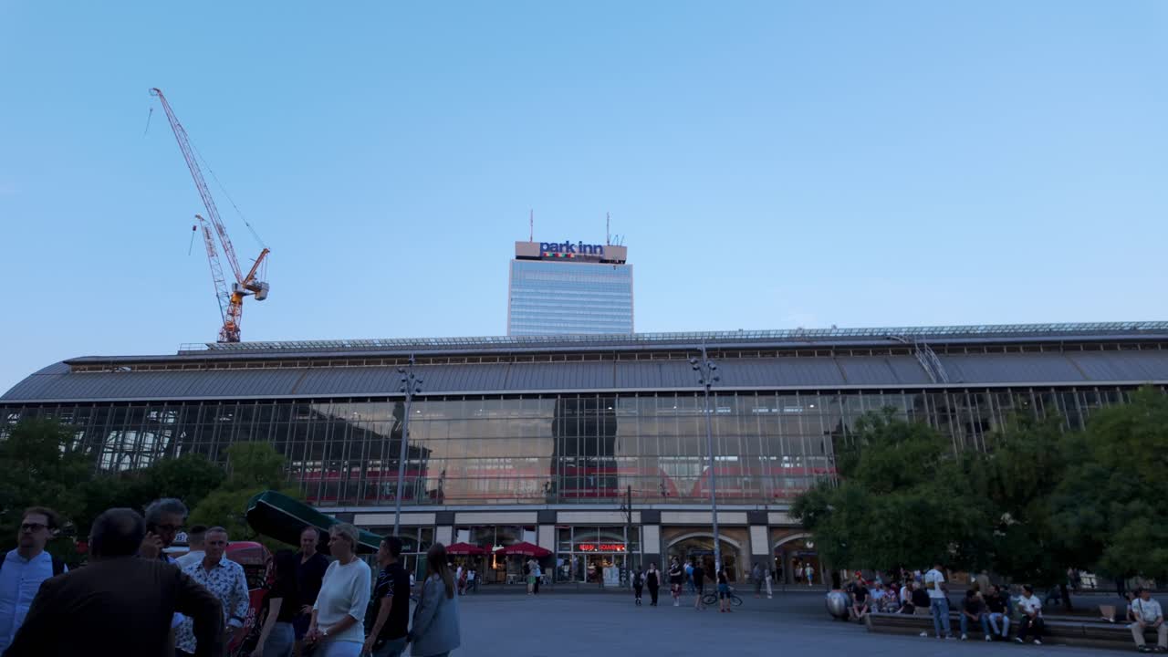 A wide-angle video shot during IFA in Berlin, featuring people walking in front of a large glass building with a tilt-up camera movement, revealing the Park Inn hotel and construction cranes above