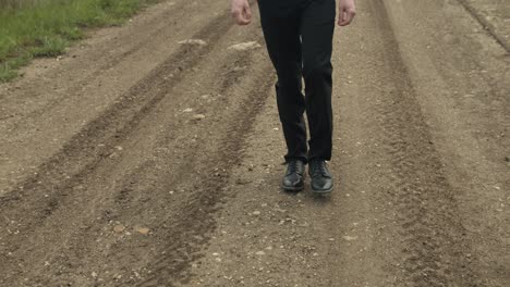 A-person-walks-steadily-down-a-gravel-road-marked-by-tire-tracks,-surrounded-by-greenery