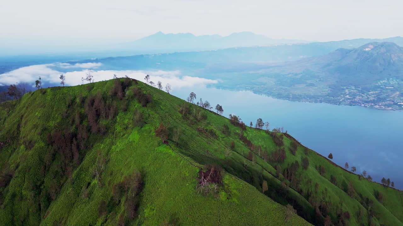 Drone footage captures the lush green landscape of Trunyan Hill in the soft early morning light with stunning views of Mount Batur and the calm waters of Lake Batur in the background.