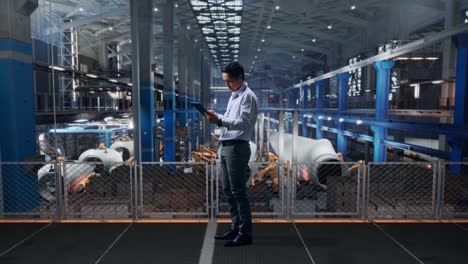 full body side view of an asian male professional worker standing with his tablet at the center of the wind turbine factory, working continuously with his tablet