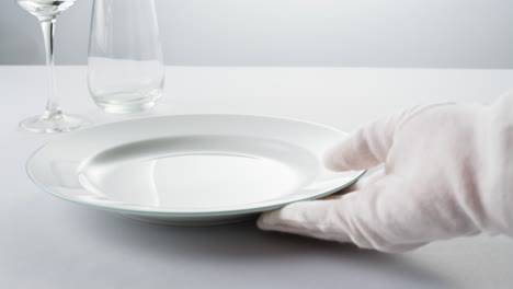 minimalist table setting with white plate, fork, and knife arranged neatly, and a gloved hand adjusting cutlery.