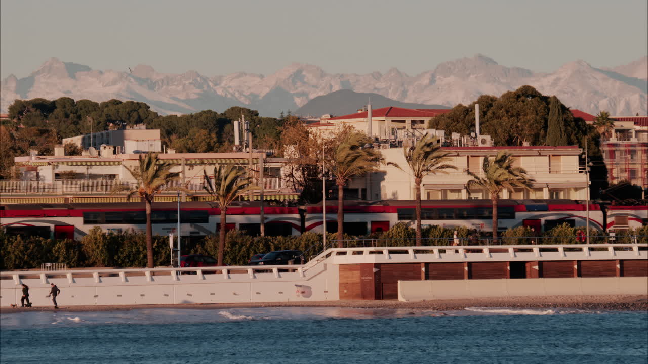 Antibes, France - November 14, 2024: Train moving in front of buildings and palm trees on the shore with a view of the mountains in the background