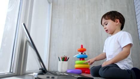 baby boy sits on the floor in front of laptop. toddler presses the keys on the keyboard. low angle view.
