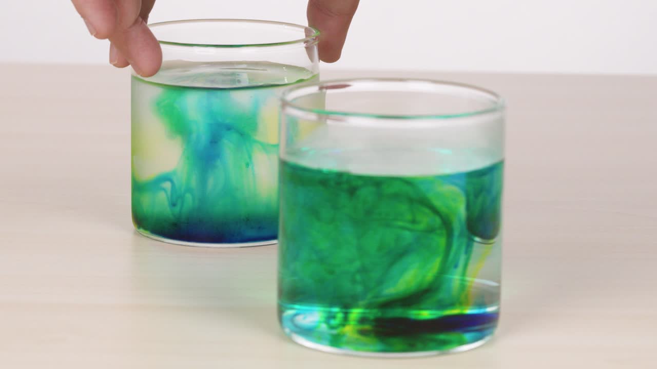 Hand demonstrates food coloring diffusion in hot and cold water, side-by-side, under soft lighting
