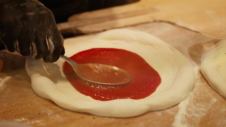 Close-up of gloved hands spreading red tomato sauce over fresh pizza dough in circular motions.