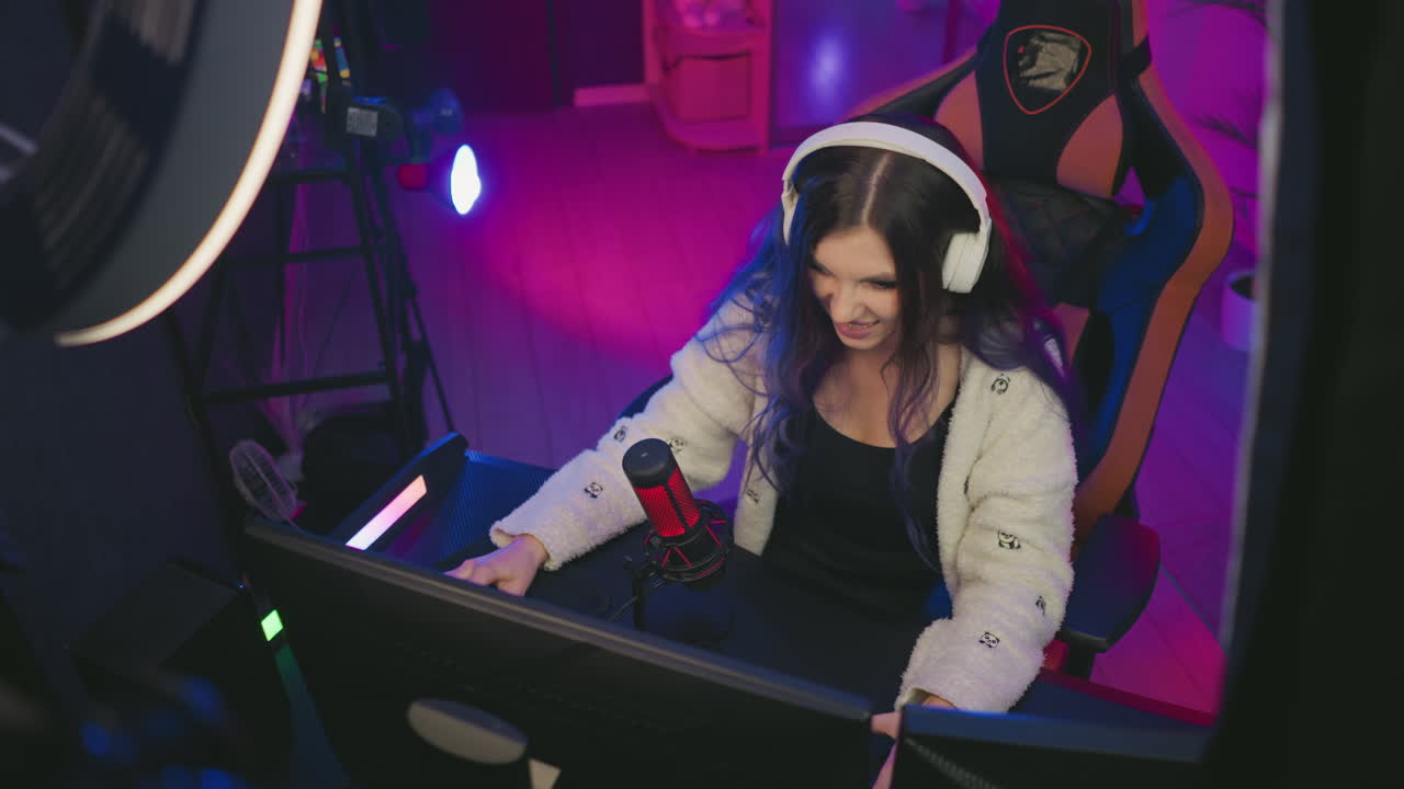 aerial view of young lady with long hair wearing headset seated at gaming desk using keyboard while playing video game surrounded by glowing neon lights, red microphone, and modern equipment