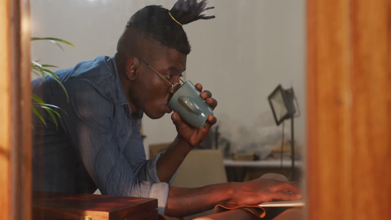 Artigiano afroamericano pensieroso con gli occhiali che beve caffè in un laboratorio di pelletteria
