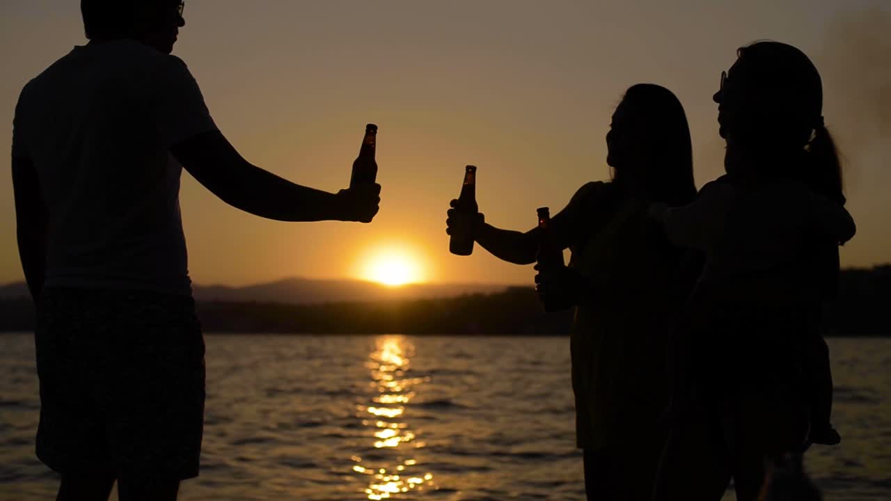 Group of young latin friends toasting with beer bottles standing on a floating dock on a lake at sunset