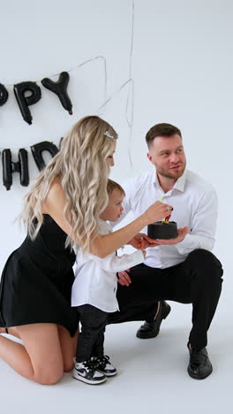 father is holding a little black cake with mickey mouse in front of his baby boy. mother is putting a candle into the cake. studio footage. vertical video.