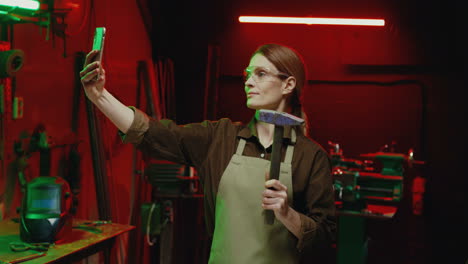 Caucasian-welder-woman-in-goggles-and-holding-hammer-making-a-selfie-with-smartphone-in-metal-workshop