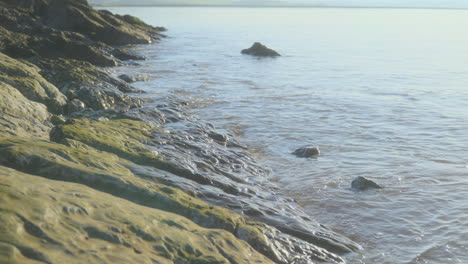 Sea-gently-breaking-against-smooth-rocks-in-early-morning-autumn-sunlight-with-slow-pan-upwards