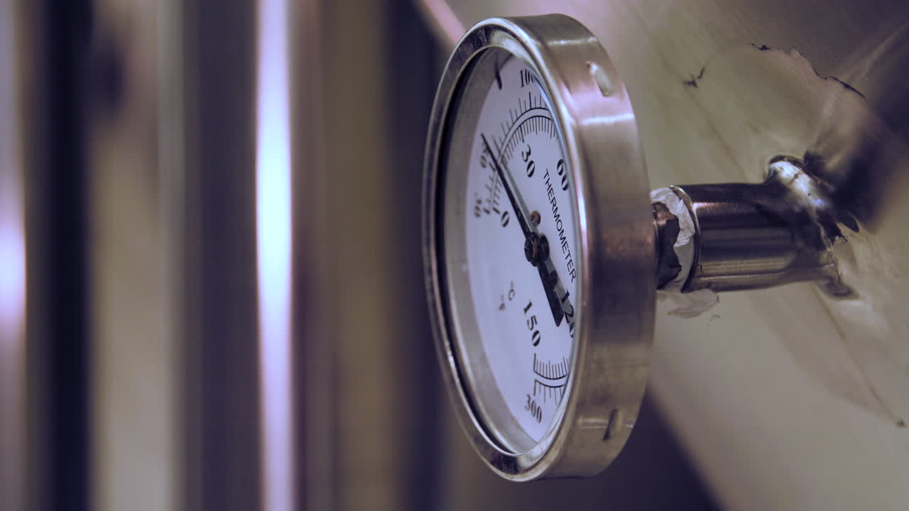 Rack focus from a blurry view to a close-up of a temperature gauge mounted on a stainless steel fermentation tank in a brewery