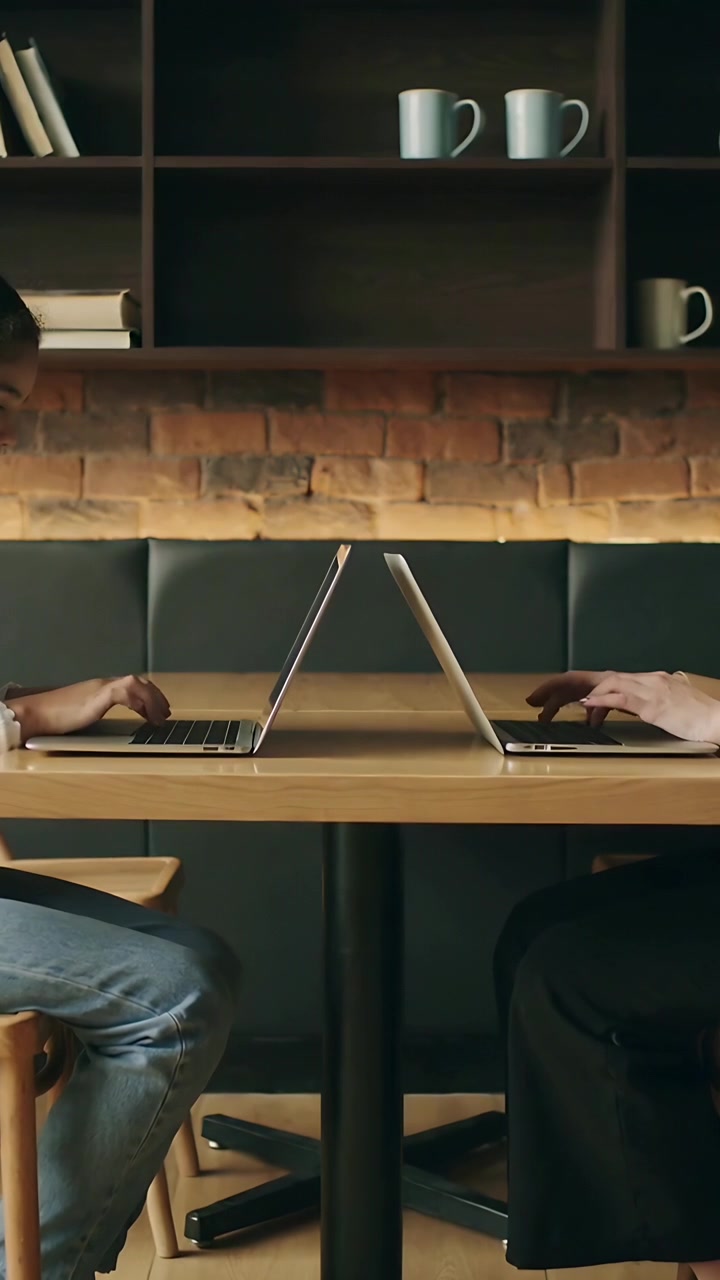 Vertical video: Typing 2 adults at cafe table, open laptops leading to work in denim and dark pants