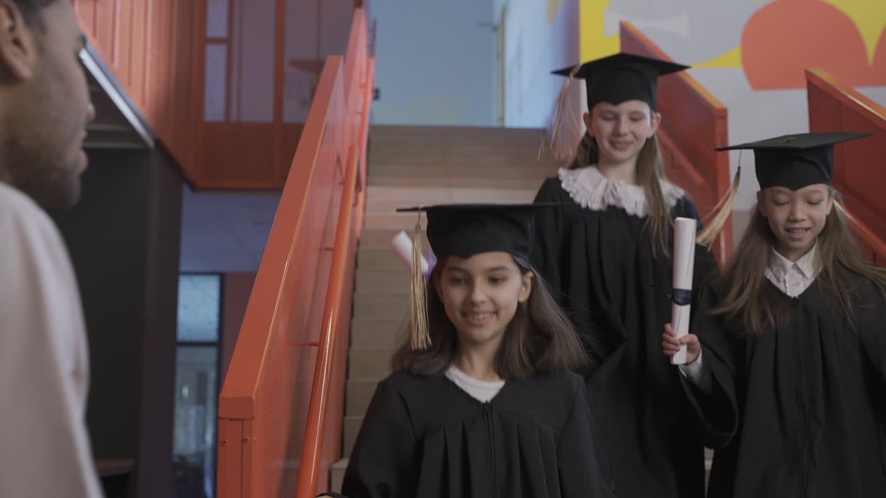 группа дошкольников в шапочках и мантиях бежит по лестнице, держа в руках дипломы об окончании школы, один из них обнимает учительницу