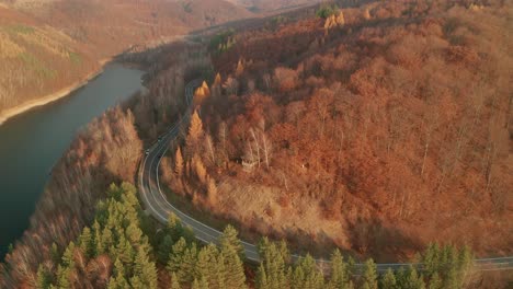 an aerial ascending drone view of a car driving on a winding mountain road alongside a lake in late autumn