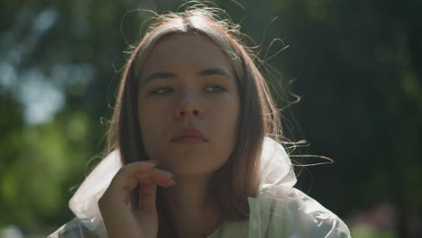 close-up of young woman in translucent raincoat thoughtfully lowering her hood and resting with eyes closed as sunlight gently filters through surrounding greenery