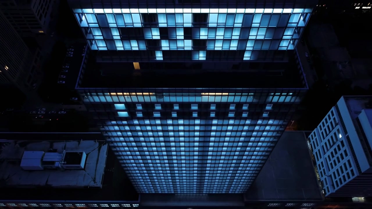 Vista noturna aérea de uma alta torre de escritórios centralizada no quadro, com luzes azuis brilhantes nas janelas formando um padrão geométrico denso em toda a fachada
