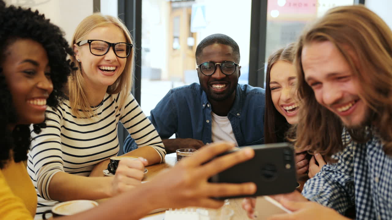 vista de primer plano de un grupo multiétnico de amigos hablando y viendo un video en un teléfono inteligente sentado en una mesa en un café