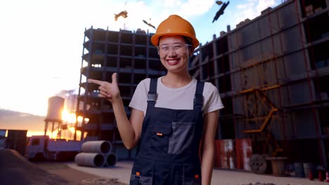 trabajadora asiática con gafas de protección y casco de seguridad sonriendo y señalando hacia un lado mientras está de pie en el sitio de construcción