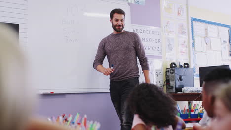 male teacher standing at whiteboard asking elementary pupils question in school classroom