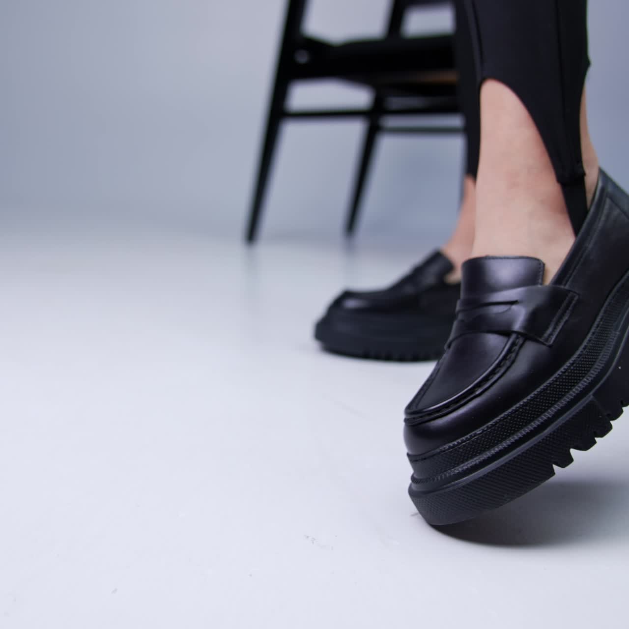 Model wearing black leggings and black shoes shows her footwear to camera. Black chair at backdrop. White studio background