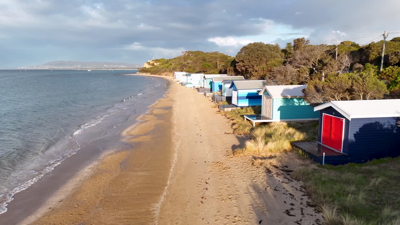 Drone glides above sandy beach, vibrant bathing boxes, and calm sea under soft daylight