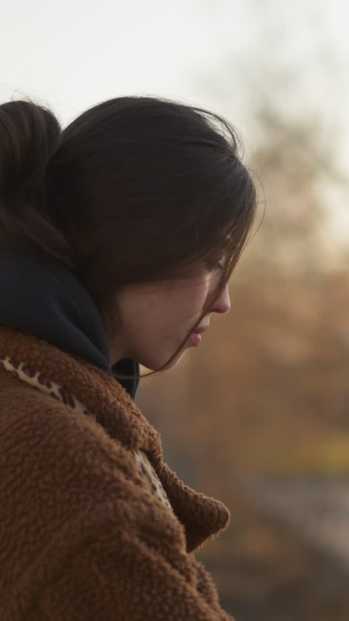 일몰에 공원을 걸어가는 슬픈 소녀의 클로즈업, 갈색 코트와 후디를 입고, 그녀의 얼굴은 머리카락에 의해 부분적으로 가려져 있으며, 부드럽고 희미한 빛에 대해 조용함을 반영합니다.