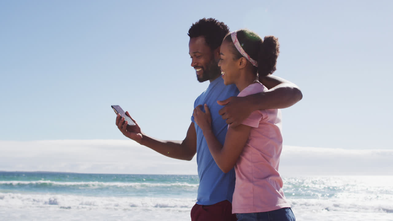 una pareja afroamericana sonriendo tomando una selfie con su teléfono inteligente en la playa