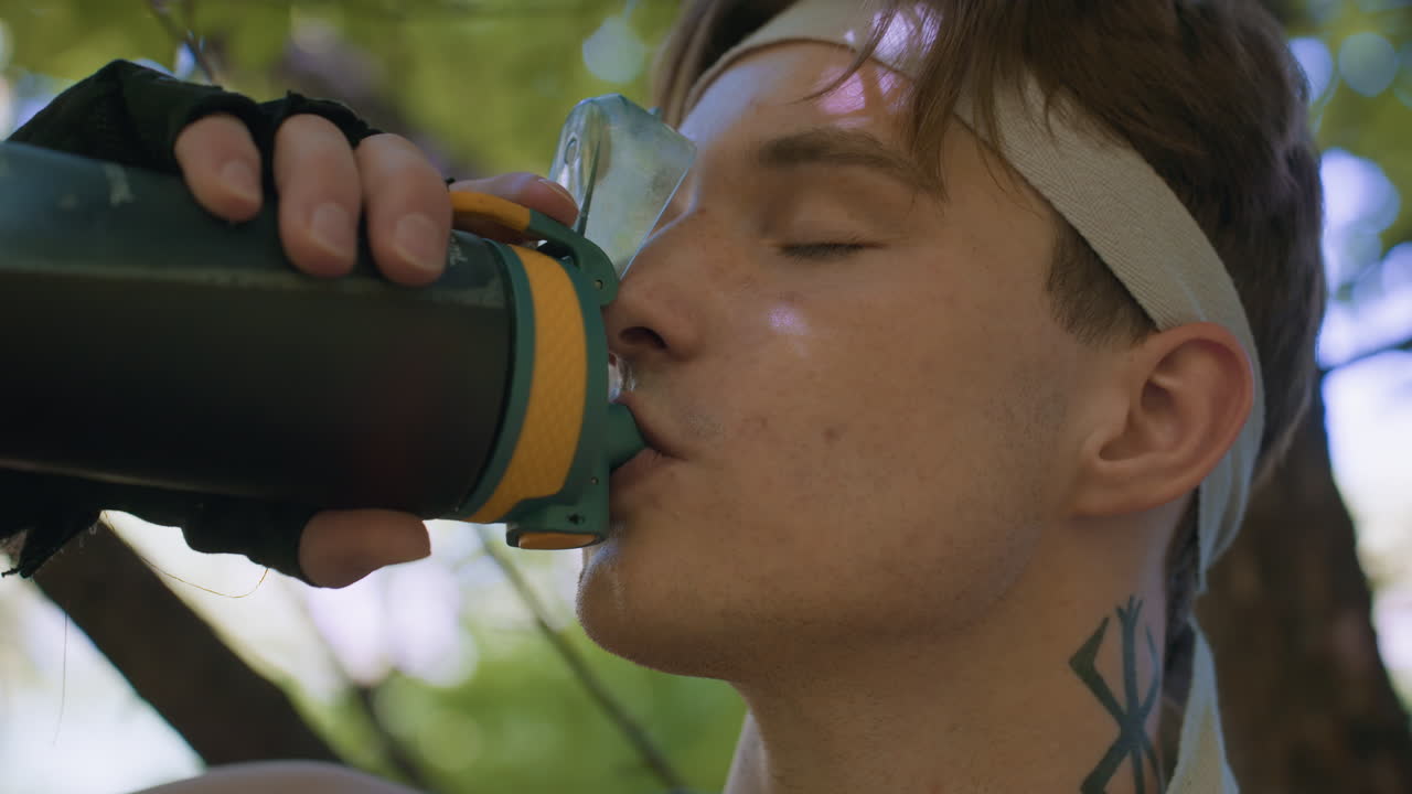 head view tourist wearing headband and glove drinking from water bottle passionately with mouth pressed to spout under leafy forest canopy, eyes closed amid dappled sunlight immersion