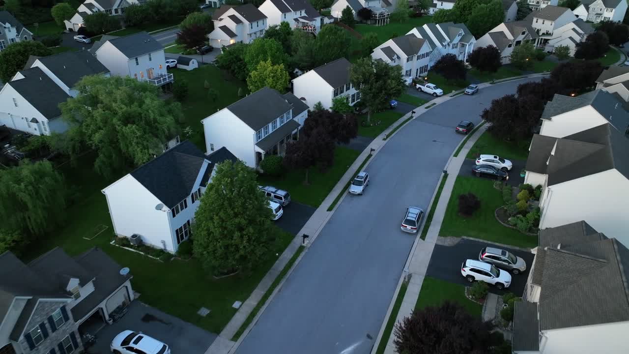 Aerial flyover american housing area with white facade and Grey roof. Porch and two-story homes with parking cars in summer. Dusk scene in the evening. Quiet suburb district with upscale buildings.
