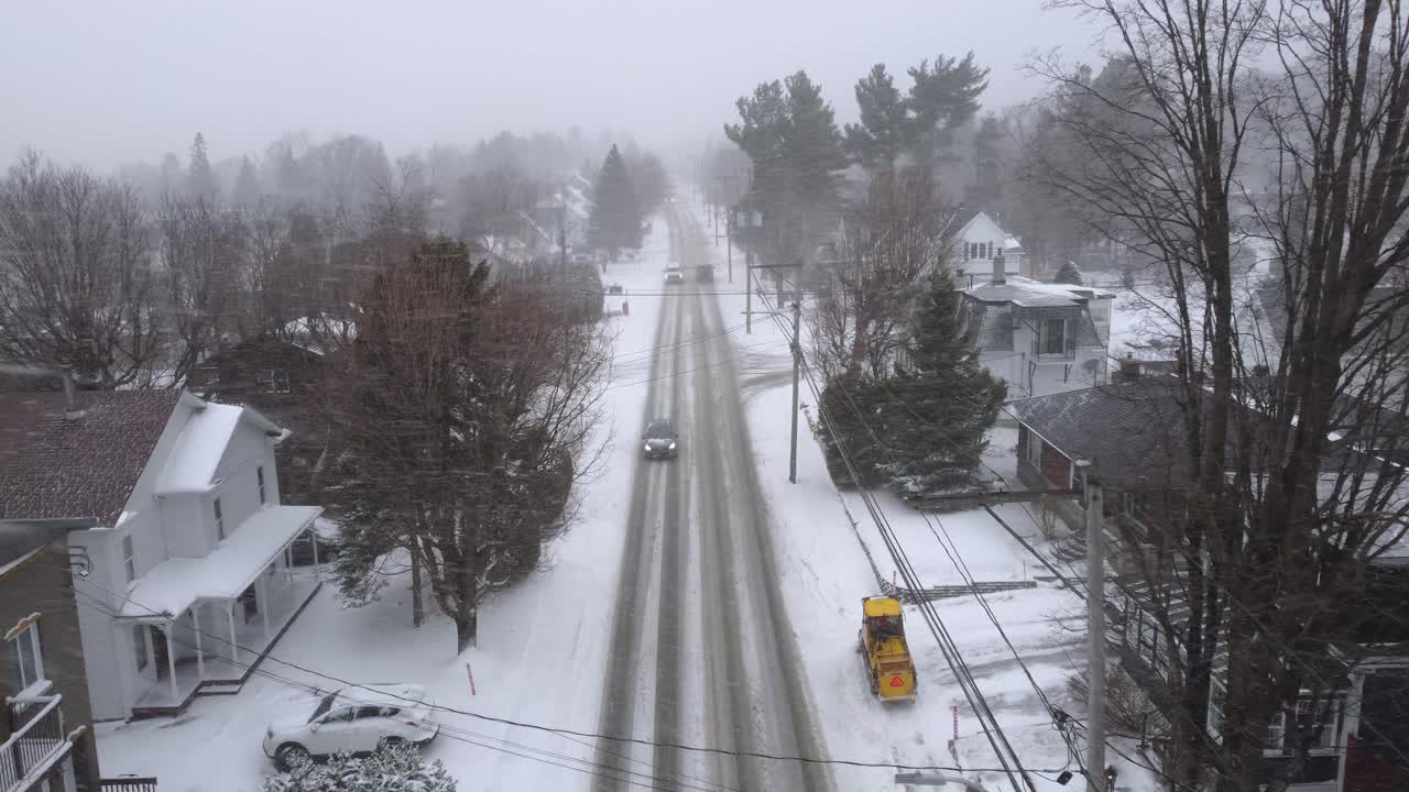 Snowplow clearing a snowy road in a residential area of Orford, Quebec, Canada, during a winter snowstorm, with snow covered houses and trees, drone descending shot