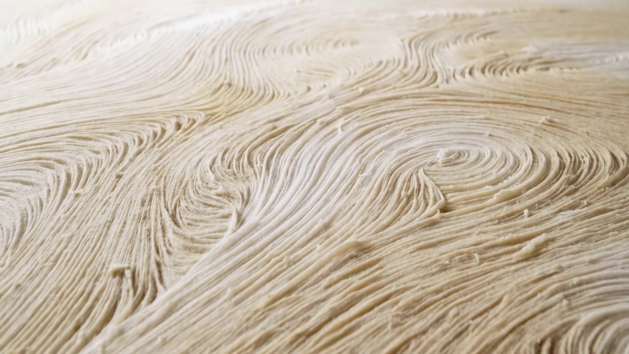 Vista macro obliqua dal basso di impasto sfogliato crudo steso su una superficie di preparazione che mostra spirali concentriche e pronunciate creste stratificate