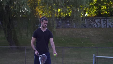 Hombre-Caucásico-De-Pelo-Largo-Blanco-Rebota-Y-Sirve-Una-Pelota-De-Tenis-Frente-A-Una-Pared-Pintada,-Parque-Comunitario-De-Hastings