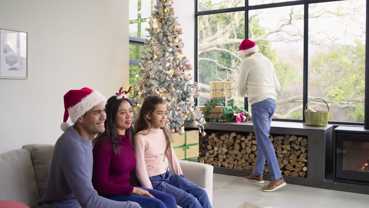 Senior man wearing Santa hat by Christmas tree selecting green gift, handing to family, copy space