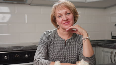 baker sits with arms crossed, lifts hand to cheek while raising head from looking at beautifully decorated cupcakes on table, expressing joy and pride in peaceful kitchen environment with lighting