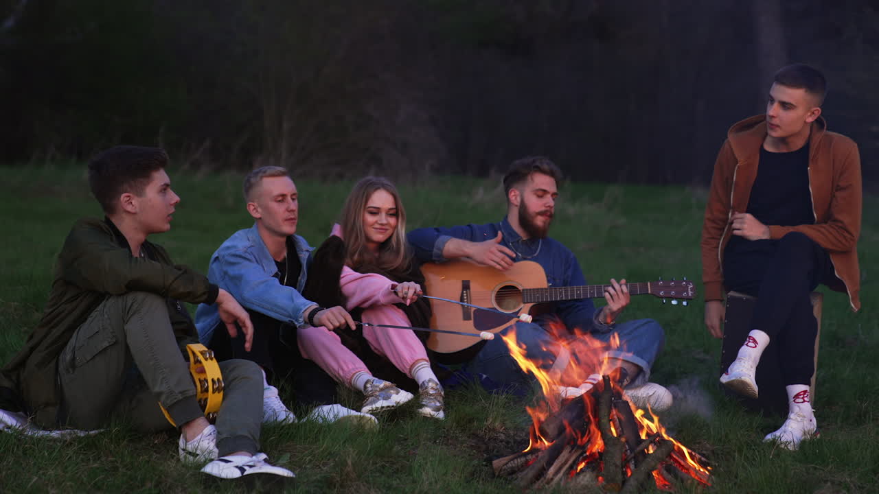 Friendly company sitting in front of fire and singing songs along with guitar. Dating couple making roast marshmallows.