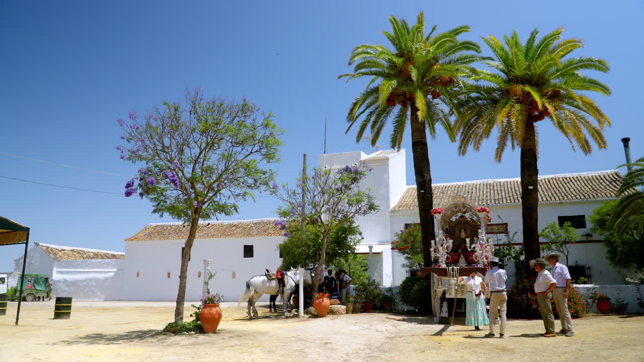 Un tradizionale cortile del Rocío sotto palme con un altare decorato e persone riunite sotto il caldo sole andaluso durante la stagione del pellegrinaggio