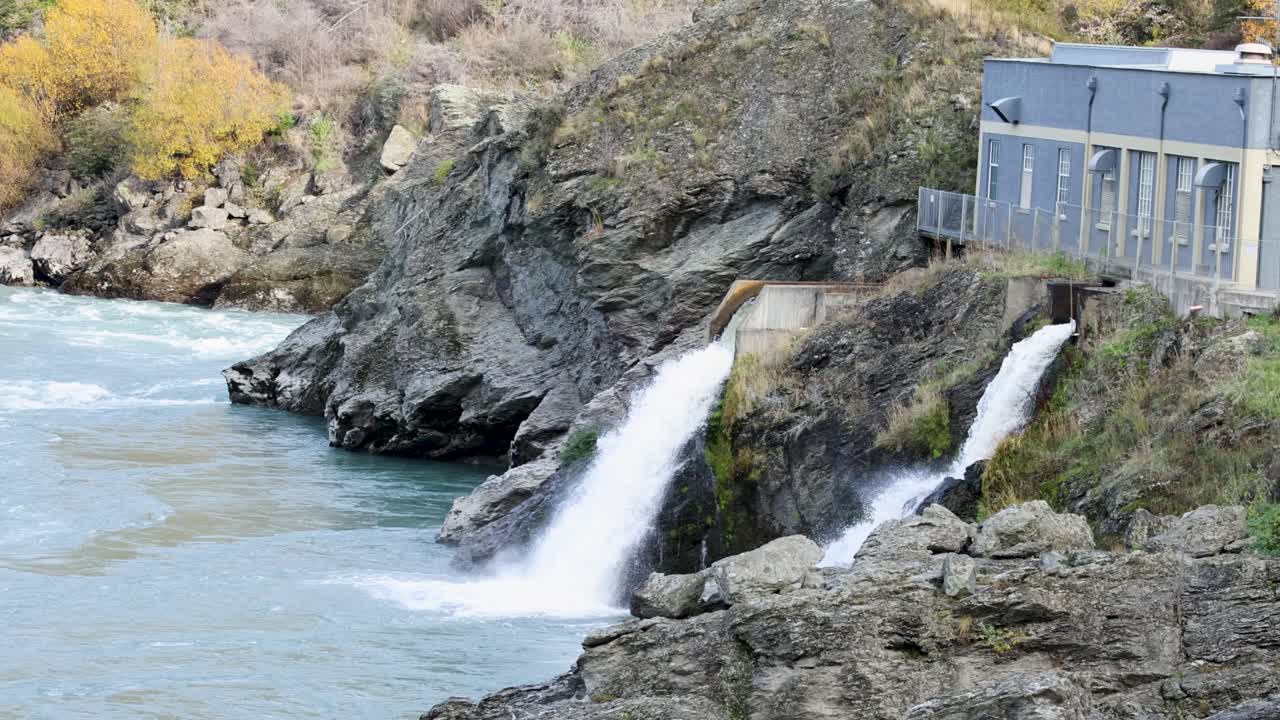 Water flows through a hydroelectric station on Kawarau River, surrounded by rocky terrain and autumn foliage in Queenstown, New Zealand