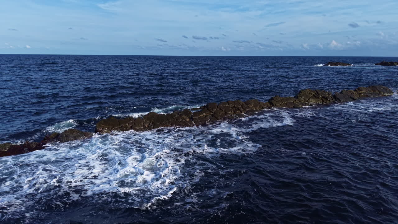 Waves crashing on rocky shoreline under a vast blue sky at midday, capturing nature's raw beauty and serenity in a coastal landscape