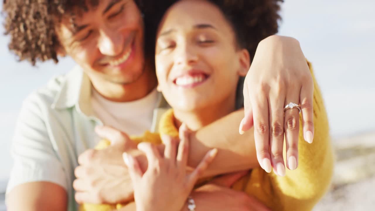 Ritratto di coppia afroamericana che sorride e mostra l'anello sulle rocce vicino al mare.
