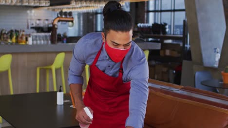 mixed race male cafe worker wearing face mask disinfecting table