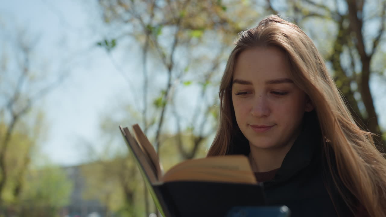Joven leyendo, estudiante absorta en un libro al aire libre, joven caucásica concentrada leyendo bajo el sol en un parque, joven lectora disfrutando del estudio en un banco de parque soleado al aire libre