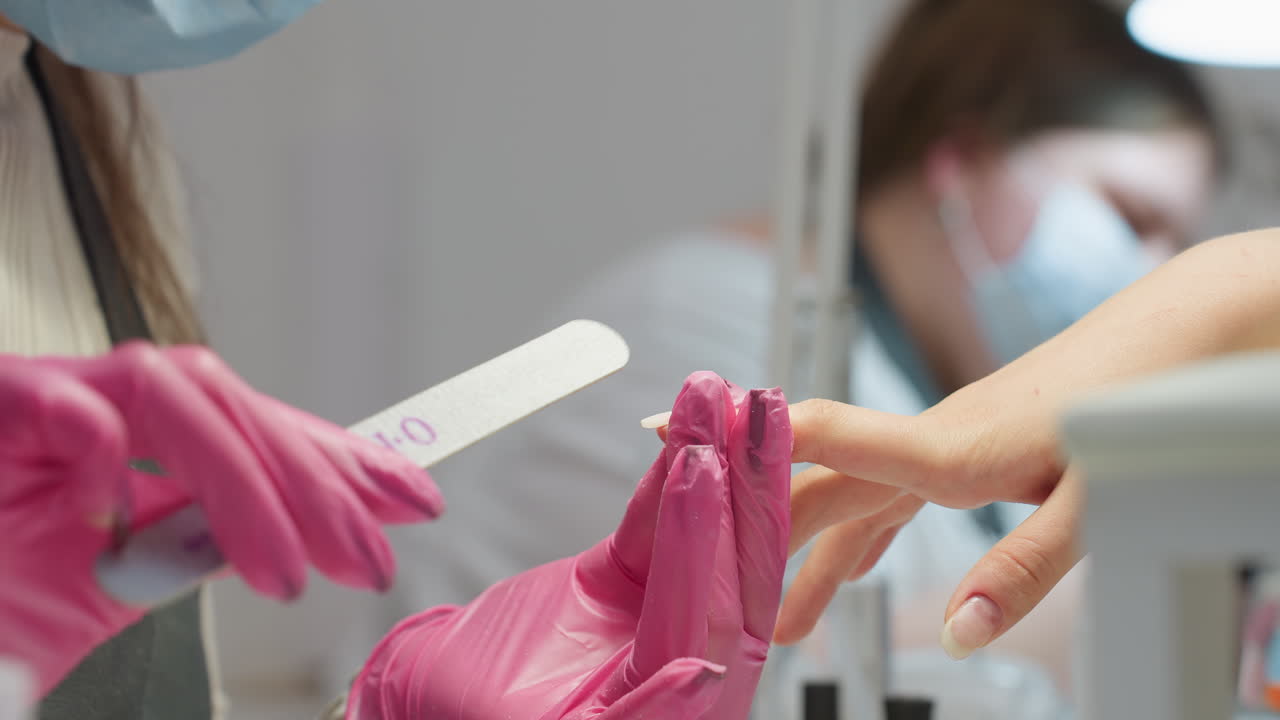 Close-up of nail technician wearing pink gloves precisely filing client's nail with second masked technician slightly blurred in background, showcasing focus and care in manicure service at busy salon